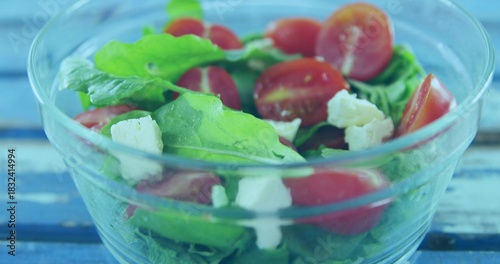 Showing clear glass salad bowl on painted blue table, with arugula, halved tomatoes, crumbled feta