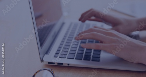 Typing woman's hands pressing keys on laptop keyboard at wooden desk, with eyeglasses nearby