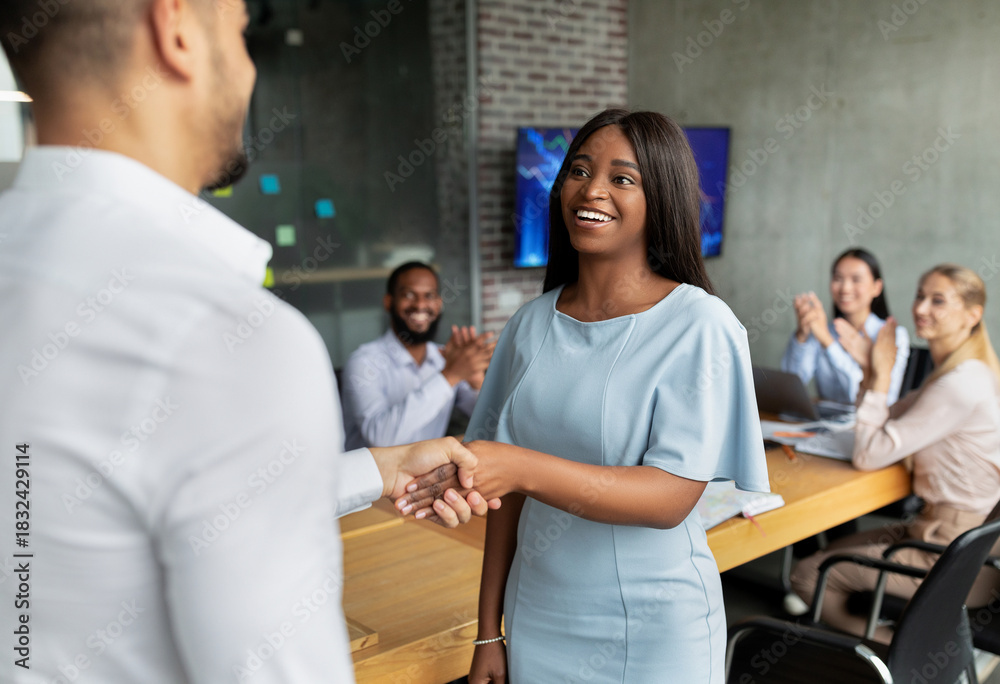 Obraz premium In a corporate meeting room, a male boss shakes hands with an African American female employee while colleagues applaud. This welcoming moment marks her first day in the team.