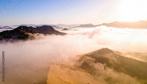 Aerial view of mountains partially obscured by a layer of clouds, illuminated by the warm light of sunrise.