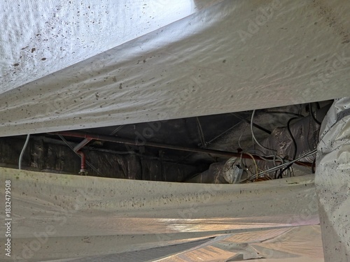 View inside a building crawl space showing a white vapor barrier covering the ground and utility pipes/wires overhead.