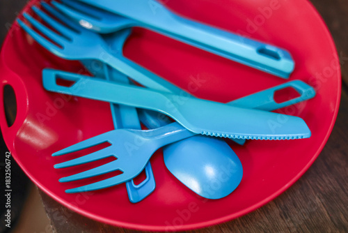 Close up of blue plastic cutlery including fork, spoon, and knife inside red bowl. These reusable utensils are ready for picnic or camping trip, simple and colorful