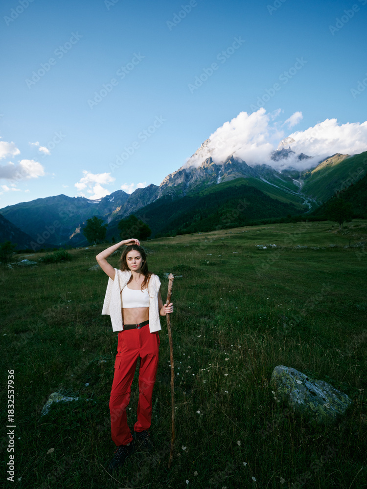 Obraz premium Hiking woman stands on a grassy alpine meadow with a trekking staff, red pants and white top, mountains in the background and a clear sky for a serene outdoor adventure