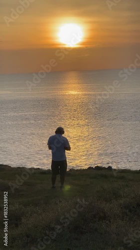 Rear view of a man on a cliff wearing headphones, enjoying the beautiful sunset over the ocean. Concept for relaxation, music, and travel escape.
