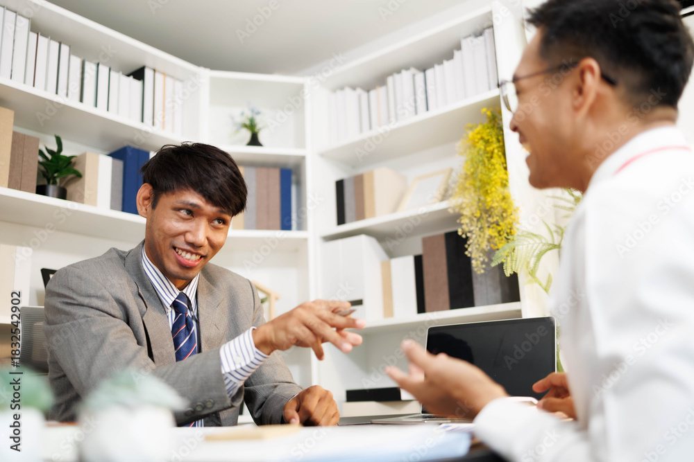 Obraz premium Two men are sitting at a desk, one of them is holding a piece of paper. They are having a conversation, and it seems like they are discussing something important. The atmosphere is tense 