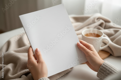 Woman holding blank brochure with hot drink and cozy blanket