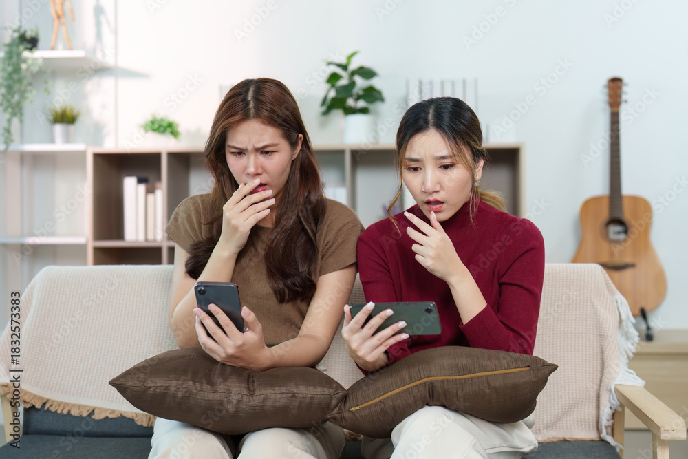 © Natee Meepian - Surprised Reactions. Two friends looking shocked while checking their phones in a cozy living room.