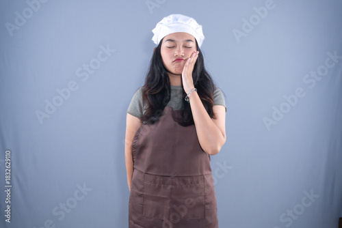 A woman in a chef's hat and brown apron holds her cheek in pain