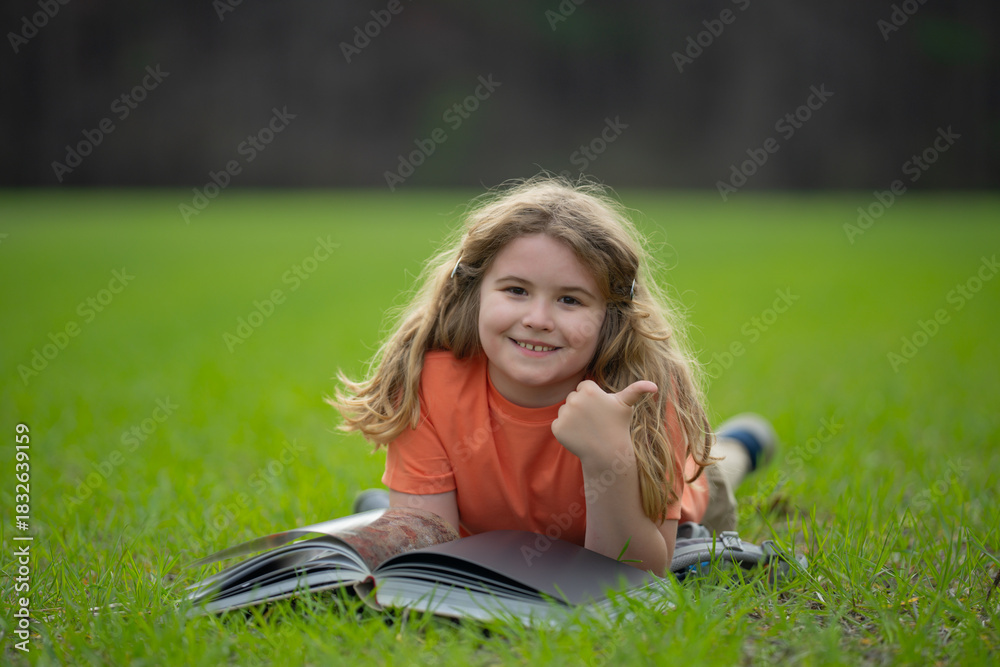 Fototapeta premium Child reading a book in the grass. Kid enjoying a book story outdoors. Child studying with book in the meadow on a sunny day. Learning in nature. Schoolboy reading book in the park. Outdoor learning.