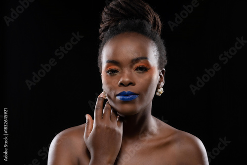 Close-up beautiful portrait black woman updo hairstyle showcasing creative makeup with blue lips and orange eyelid against at studio shot on black background.