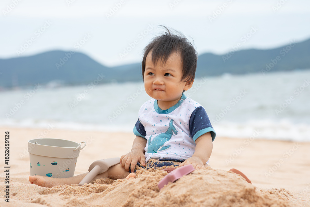 Fototapeta premium Asian family baby sitting on sandy beach with bucket and shovel smiling at seaside