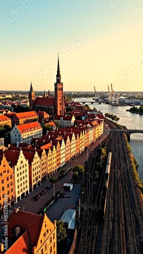 Aerial view of a city with a tall church and red roofed buildings alongside train tracks and water at sunset