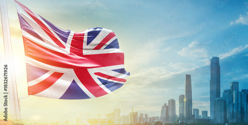 Obraz premium United Kingdom flag wavering in beautiful clouds with building skyline. The flag with drone shot in beautiful sky. UK national flag for independence day.