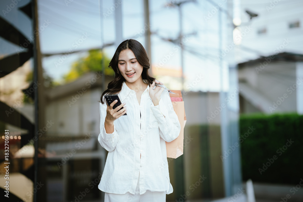 Fototapeta premium An Asian woman happily talking on the phone while holding shopping bags outside a modern building, capturing the joyful lifestyle of a young Asian woman on a shopping day.