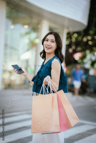 An Asian woman happily looking at her phone while carrying shopping bags outside a modern building, capturing the cheerful lifestyle of a young Asian girl enjoying her shopping day.