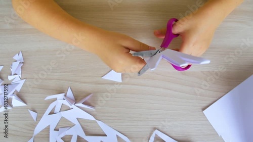 A child creating paper white snowflakes at a table at home. A child cuts patterns on paper with scissors. Decoration for the winter holiday.