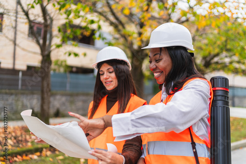 Two women engineers wearing hard hats and safety vests discussing a blueprint outdoors, collaborating on a project plan