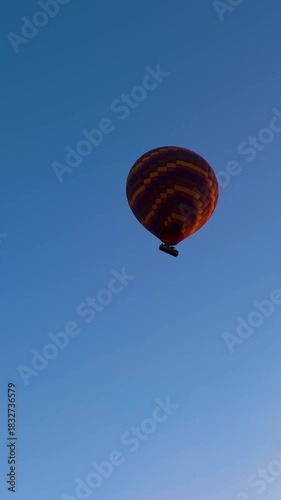 Vertical video. Goreme, Nevsehir, Turkey. Single large checkered hot air balloon with yellow red orange purple colors and basket with tourists in morning blue sky. Aerial View. Rich colors