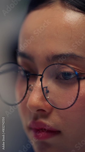 An extreme close-up of a young Asian woman wearing round glasses and concentrating intensely while working late at night in front of a computer screen.