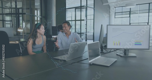 Talking pair wearing sleeveless top and blue shirt at table in office with silver laptops, monitor