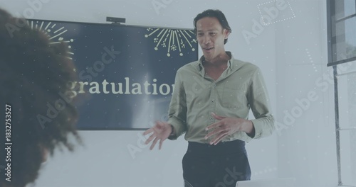 Speaking presenter in green shirt and dark pants in meeting room, with display showing gratulation