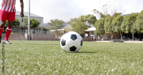 Rolling soccer ball on trimmed grass field, player in red-striped jersey approaching left