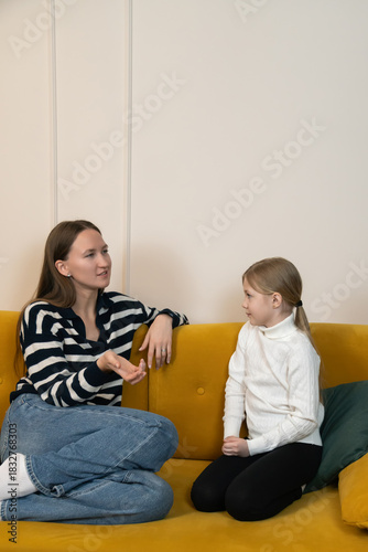 Mother and daughter enjoying a happy conversation and bonding time, smiling and sharing moments while sitting casually on a yellow sofa in a comfortable home environment. Vertical photo