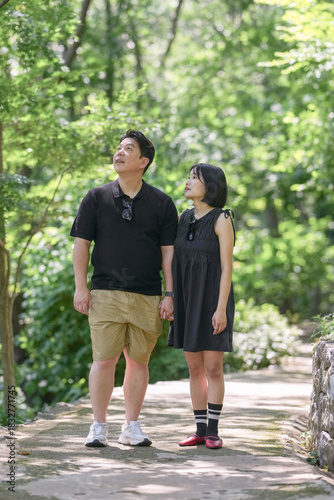 A man and a woman are standing on a bridge, looking at something in the distance. The man is wearing a black shirt and khaki shorts, while the woman is wearing a black dress