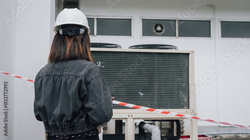 An engineer wearing a safety helmet examines an air conditioning unit at a commercial building. The area is marked off with caution tape to indicate restricted access