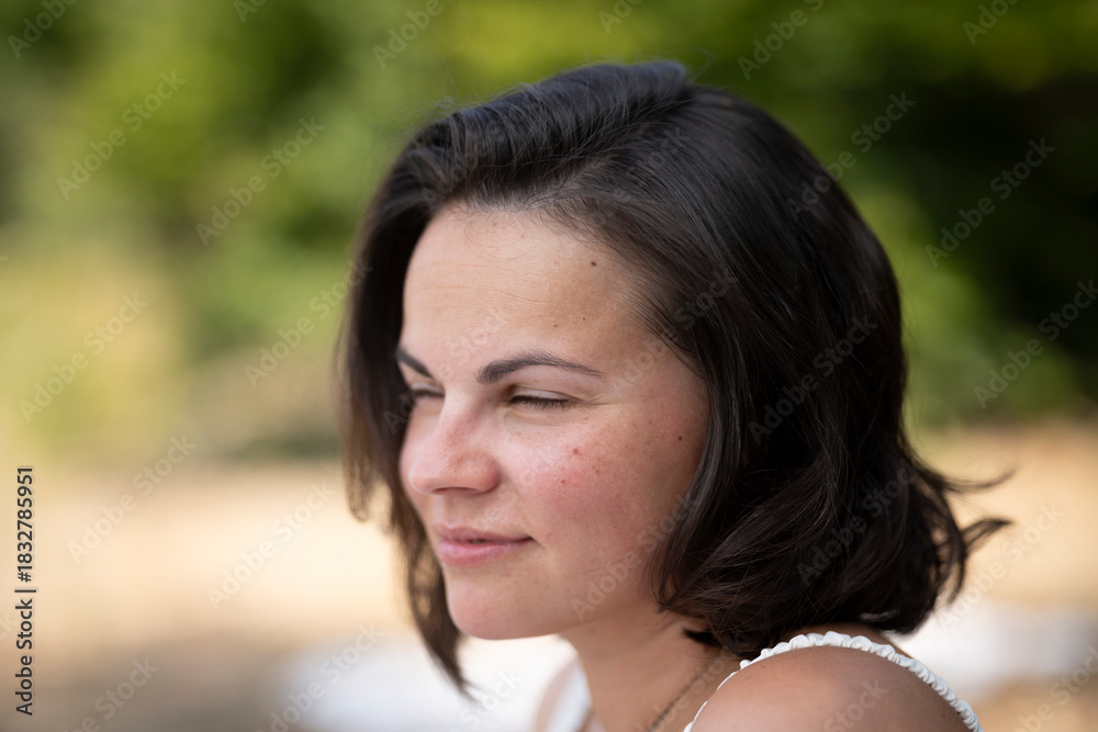 Naklejka premium Beautiful dark-haired woman smiling with squinted eyes sitting on summer beach, looking into the distance, emotional portrait with copy space and blurred background