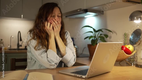 Exhausted and sleepy female freelancer struggling with deadlines while working on her laptop late at night in the kitchen, trying to stay awake and focused on her remote work from home