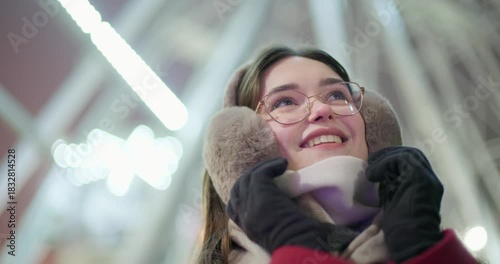 A young beautiful girl in a red coat walks in the evening at the New Year's Christmas fair against the backdrop of a Ferris wheel among lights and garlands and admires the decorations