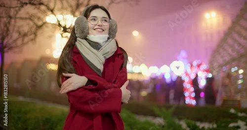 A young beautiful girl in a red coat walks in the evening at the European Christmas Market against the backdrop of illuminations and garlands.