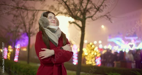 A young beautiful girl in a red coat walks in the evening at the European Christmas Market against the backdrop of illuminations and garlands.