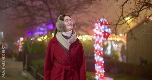 A young beautiful girl in a red coat walks in the evening at the European Christmas Market against the backdrop of illuminations and garlands.