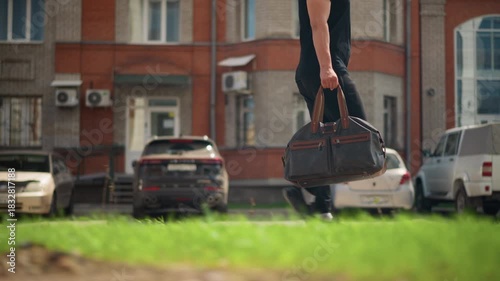 man with leather bag, closeup of man walking outdoors, closeup of man carrying leather bag on sunny street, man walking along sunny pavement carrying leather bag beside parked cars in city