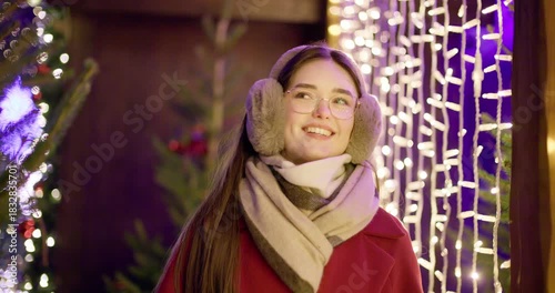 A young beautiful girl in a red coat walks in the evening at the European Christmas Market against the backdrop of illuminations and garlands.