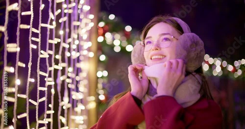 A young beautiful girl in a red coat walks in the evening at the European Christmas Market against the backdrop of illuminations and garlands.