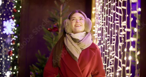 A young beautiful girl in a red coat walks in the evening at the European Christmas Market against the backdrop of illuminations and garlands.