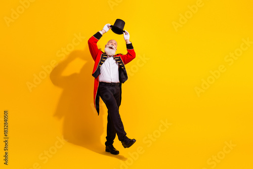 Senior magician in red coat lifts top hat against bright yellow background during a festive performance and retro style showman moment