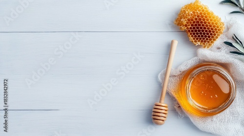 A top-down shot of a jar of honey, a honeycomb, and a honey dipper on a white wooden surface. The scene is lit with natural light.