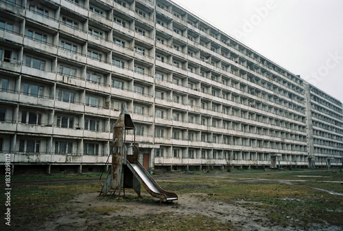 Old, abandoned playground with a rusty slide against a long, brutalist apartment building. A grim, post-soviet urban landscape on an overcast day.