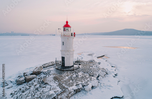 On an icy island in Vladivostok, a lighthouse stands surrounded by frozen water, with the dawn casting a calming glow.