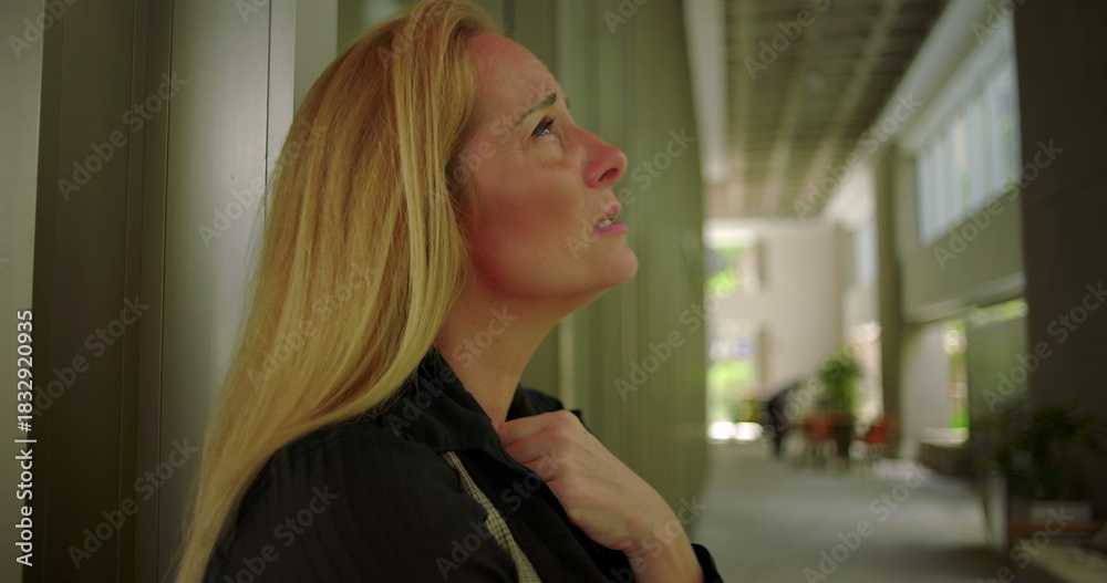 Fototapeta premium Side view of woman lowering her gaze with hand near chest in corporate corridor with modern seating and soft natural light through windows