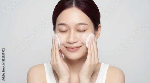 A woman with closed eyes gently cleanses her face with cotton pads, performing a relaxing skincare routine in a bright studio.