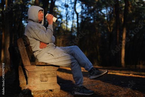 Morning sun casts golden light on a man sitting alone on a bench, enjoying a warm drink in a peaceful park surrounded by tall trees