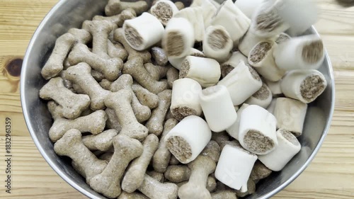Closeup of dog biscuit cylinder shape rolls being poured into a large steel bowl, filling it up next to crunchy bone shaped biscuits.