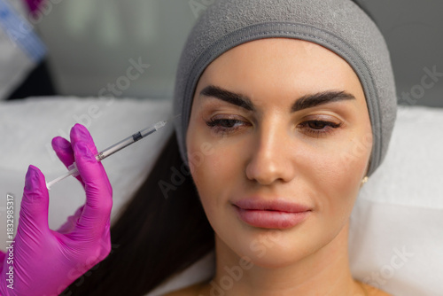 Close up of hands of young cosmetologist injecting botox in female face. She is standing and smiling. The woman is closed her eyes with relaxation