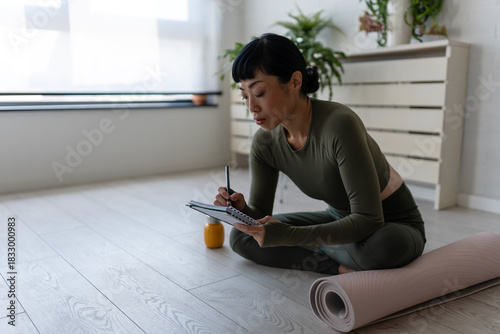 Woman in sportswear sitting on floor by yoga mat, writing in notebook and reflecting after home workout or yoga session
