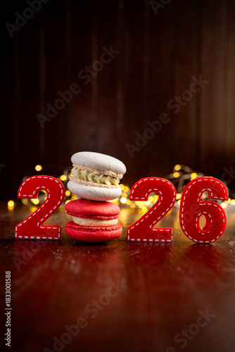 Stacked macarons and felt numbers 2026 sit on a dark wooden table. Creating a festive holiday atmosphere with blurred yellow lights in background. Symbolizing new beginnings and sweet celebrations